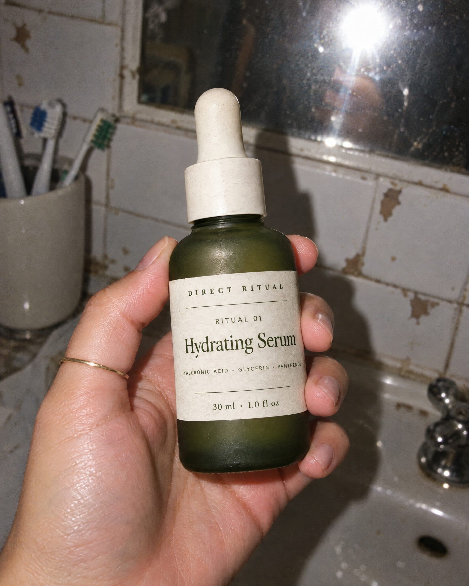A hand holding the Direct Ritual serum bottle in a bathroom, lit by flash